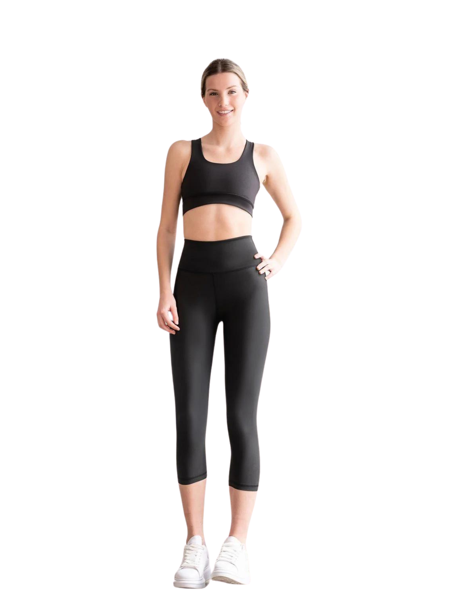 Person in Lismina 4306 Capri Leggings in Schwarz und weißen Sneakers steht lächelnd mit Händen an Hüften vor Weiß.